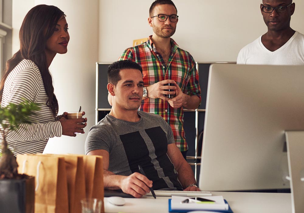 A man sitting and working on a desktop computer. A woman and two men surround him and look at the computer’s screen