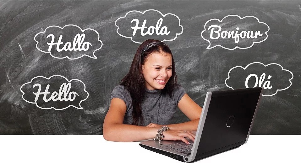 Young person sitting at a laptop with a chalkboard in the background. The chalkboard has the English word "hello" translated in four different languages.