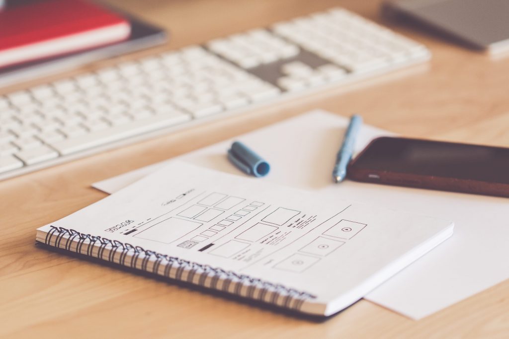 A desk with a keyboard, a spiral notebook, a pen, and a smartphone