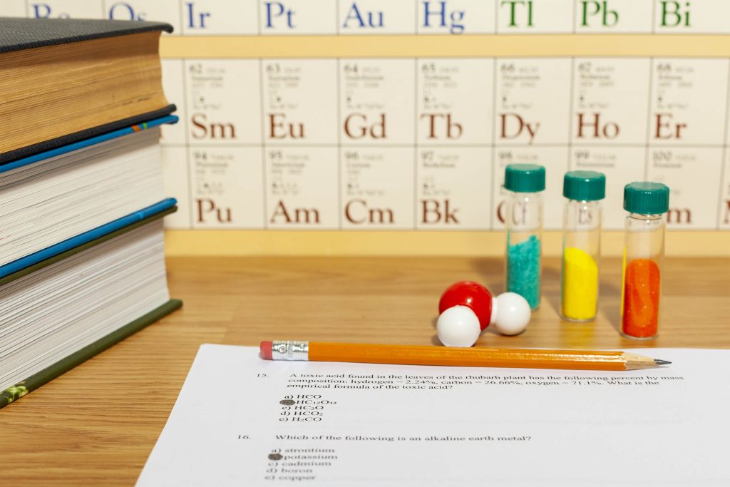 A tabletop with a sheet of paper with multiple choice questions marked. A pencil is laying on top. A periodic table chart, a molecule, powder vials, and books are on the table.