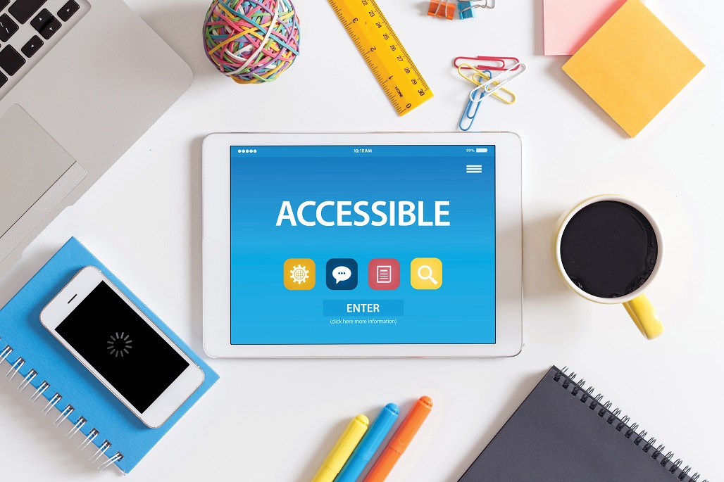 Desktop filled with a tablet that says accessible, a smartphone, a spiral notebook, pens, a ruler, paperclips, sticky notes, and a cup of coffee